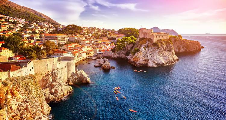 La lumière dorée du soleil sur les anciens murs de la vieille ville de Dubrovnik et la forteresse perchée sur la côte rocheuse avec des kayakistes en contrebas.