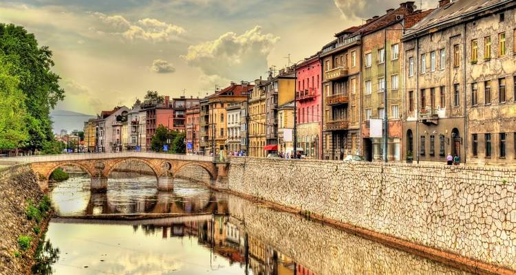 Pont en pierre et bâtiments riverains colorés reflétés dans la rivière Miljacka à Sarajevo sous un ciel dramatique.