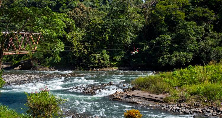 Une personne pratiquant la tyrolienne au-dessus d'une rivière à courant rapide dans une forêt tropicale dense.