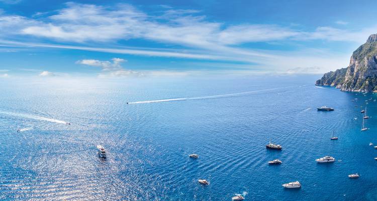 Panoramisch luchtfoto van Capri's glinsterende blauwe zee vol met jachten en steile kalkstenen rotsen onder een heldere hemel.