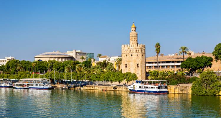 Torre del Oro and riverside promenade with tour boats reflecting on the Guadalquivir River in Seville.