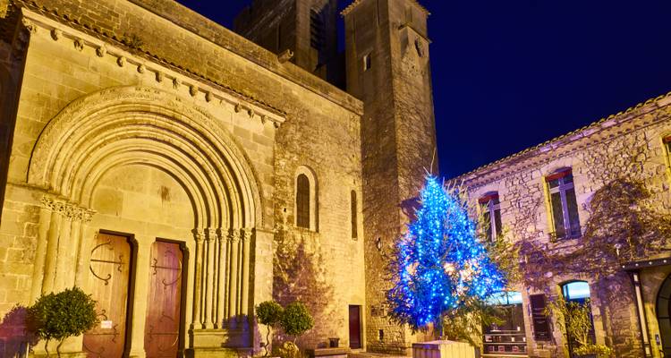 Église en pierre et clocher illuminés la nuit à côté d'un sapin de Noël éclairé en bleu dans une place médiévale.