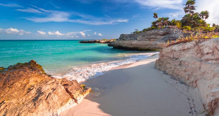 Plage tropicale immaculée avec eau turquoise, affleurements rocheux et sable blanc sous un ciel bleu.