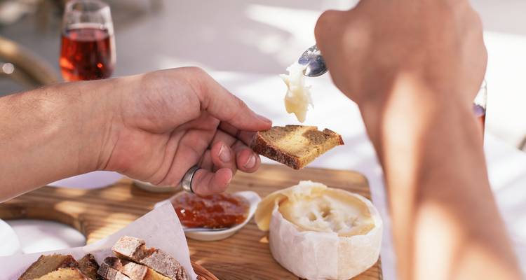 Une main étale du fromage onctueux sur du pain rustique avec de la confiture et du vin rosé à proximité.