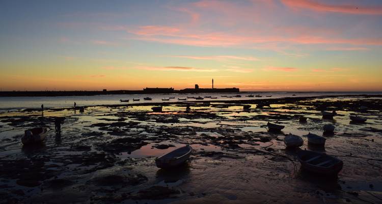Coucher de soleil coloré au-dessus des vasières à marée basse avec de petits bateaux et un phare lointain à Cadix