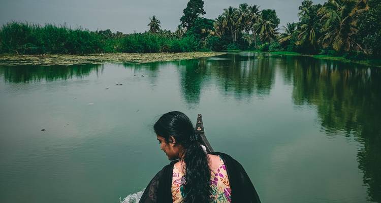 Eine Frau paddelt durch einen schmalen Kanal, der von üppigen Palmen und lotusbedecktem Wasser in den Backwaters von Kerala gesäumt wird.