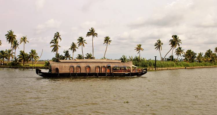 Ein Hausboot in Kerala gleitet an Reihen schiefer Palmen unter einem teilweise bewölkten Himmel vorbei.