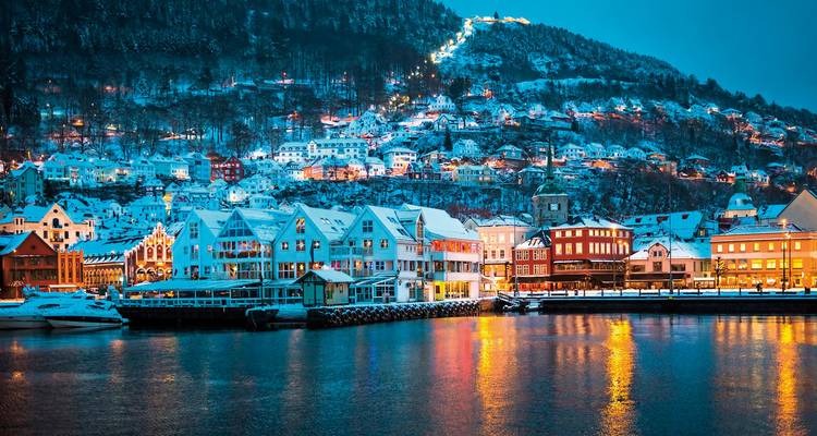 Der schneebedeckte Hafen von Bergen spiegelt farbenfrohe Häuser und Hügellichter im ruhigen Gewässer nach Einbruch der Dunkelheit wider.