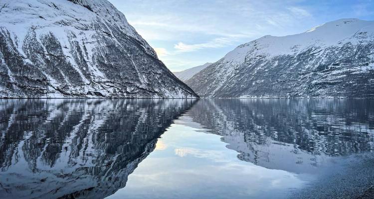 Der spiegelglatte Fjord reflektiert schneebedeckte Berge und den blauen Winterhimmel in der ruhigen Region Geirangerfjord.