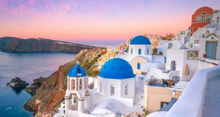 Las icónicas iglesias de cúpula azul de Oia bajando en cascada por la caldera de Santorini en un amanecer de colores pastel.