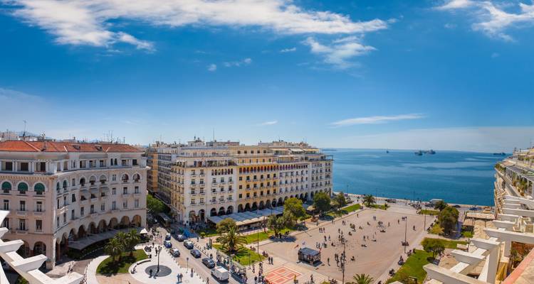 Panorámica amplia de la Plaza Aristotelous y el paseo marítimo de Tesalónica con edificios neoclásicos y multitudes paseando bajo un cielo azul brillante.