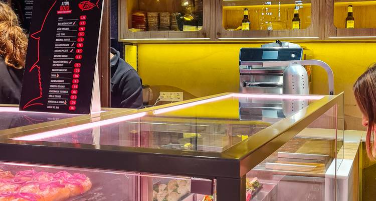 Comptoir de bar à tapas avec vitrine en verre, éclairage rose et tableau de menu tandis que les clients attendent.