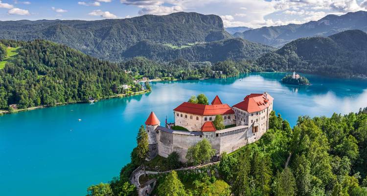 Luftaufnahme des mittelalterlichen Schlosses Bled auf einer Klippe neben einem leuchtend blauen See und alpinen Bergen.
