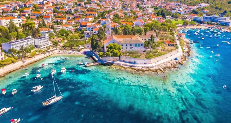 Luftaufnahme einer kroatischen Küstenstadt mit türkisem Wasser, Segelbooten und roten Dächern an einem sonnigen Tag.