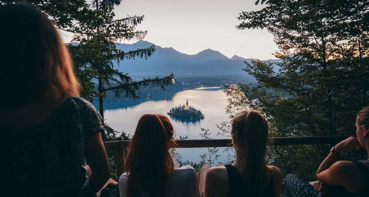 Los viajeros admiran el Lago Bled y su iglesia de isla desde un mirador boscoso al atardecer.