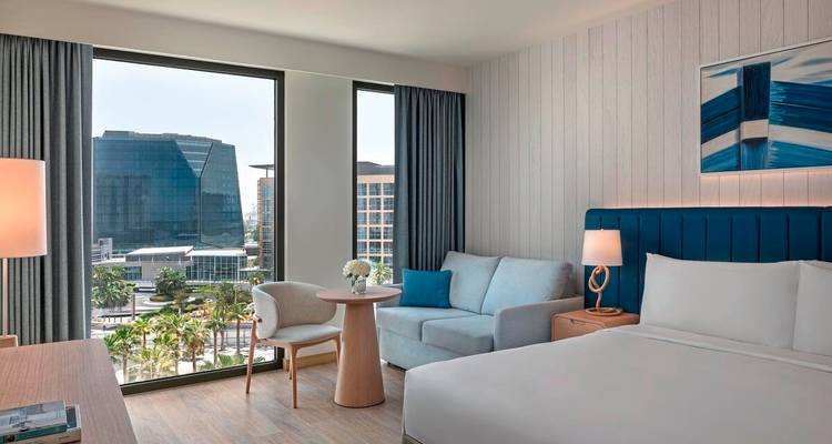 Helles modernes Hotelzimmer mit großen Fenstern mit Blick auf zeitgenössische Architektur und einer palmengesäumten Allee.