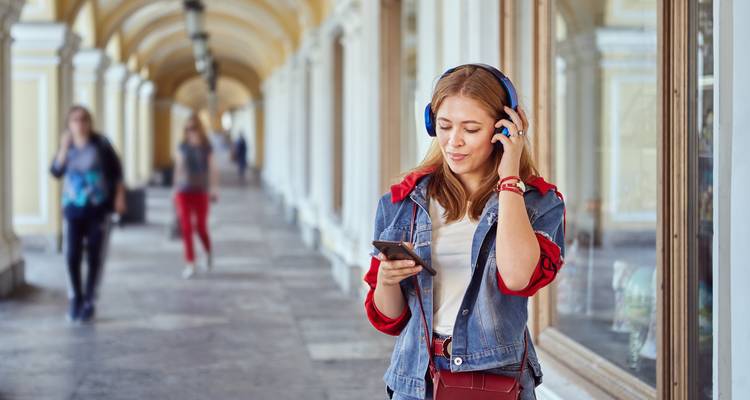Lächelnde junge Frau mit Kopfhörern nutzt ihr Telefon, während sie unter Arkaden spaziert