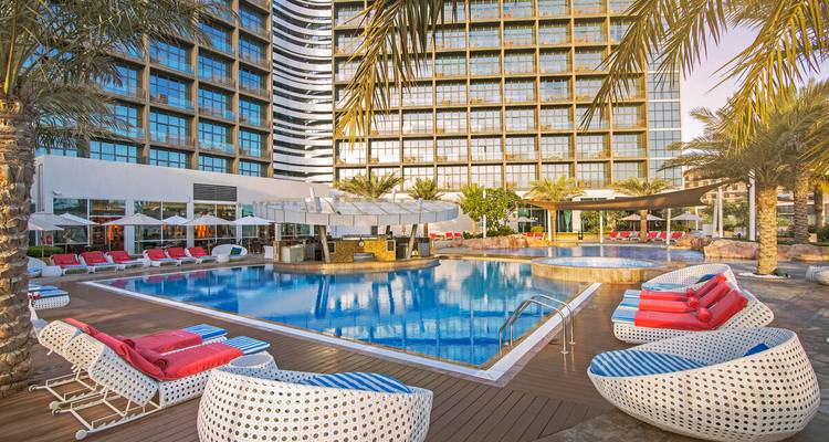 Schicke Hotelpool-Anlage mit farbenfrohen Liegestühlen, Palmen und einer Hochhaus-Glasfassade im Hintergrund.