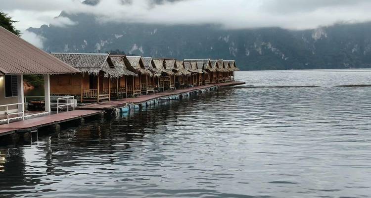 Une rangée de bungalows flottants avec toit de chaume s'étend sur un lac tranquille entouré de falaises calcaires brumeuses.