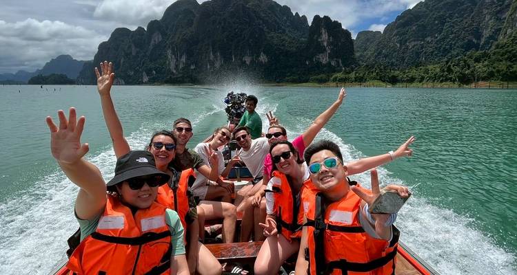 Les voyageurs souriants en gilets de sauvetage agitent la main sur un bateau à queue longue qui file à toute vitesse sur un lac d'émeraude avec des pics calcaires.