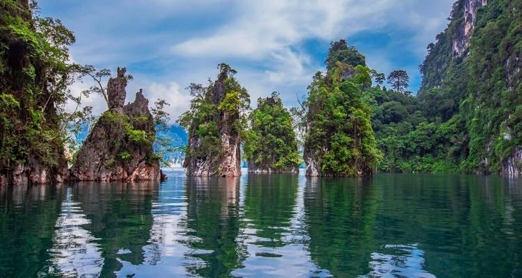 Lac calme reflétant des îles de calcaire imposantes et des falaises de forêt tropicale luxuriante sous un ciel partiellement nuageux à Khao Sok.