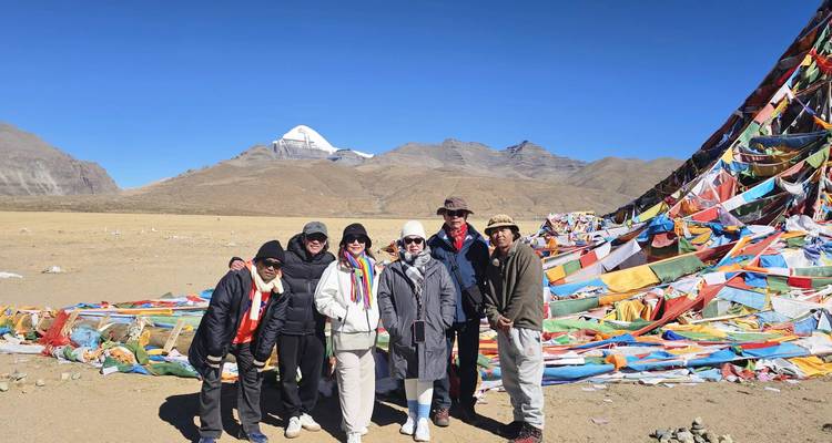 Un petit groupe se tient près de monticules de drapeaux de prière colorés avec le Mont Kailash aux sommets enneigés qui brille sous un ciel dégagé.