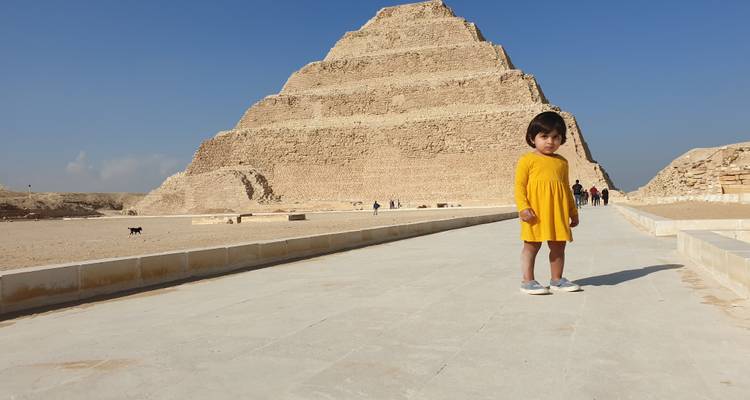 Kleines Kind in gelbem Kleid steht vor der Stufenpyramide unter blauem Himmel.