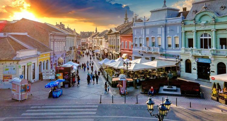 Rue piétonne animée à Novi Sad bordée de bâtiments colorés sous un ciel dramatique au coucher du soleil.