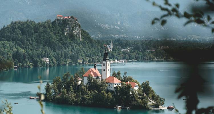 Luchtfoto van eilandkerk op Meer Bled met beboste heuvels en kasteel op verre rots.