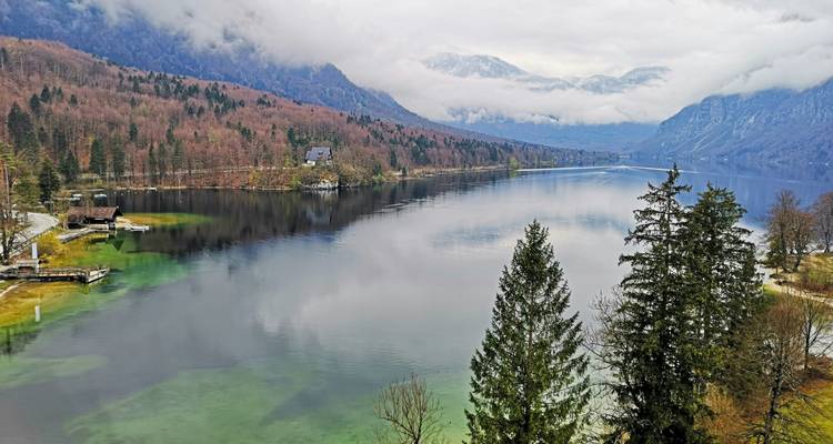 Grillig panorama van meer Bohinj met mistige bergen en herfstbossen weerspiegeld in rustig water.
