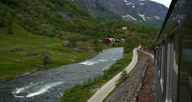 Vue depuis la fenêtre d'un train d'une rivière tumultueuse longeant des vallées verdoyantes et des plaques de neige lointaines