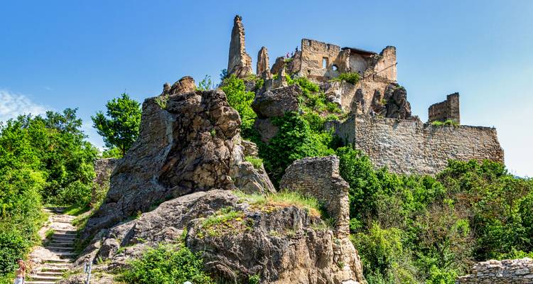 Las ruinas de piedra robusta del Castillo de Dürnstein encaramadas en una colina rocosa rodeadas de exuberante vegetación bajo un cielo azul despejado.