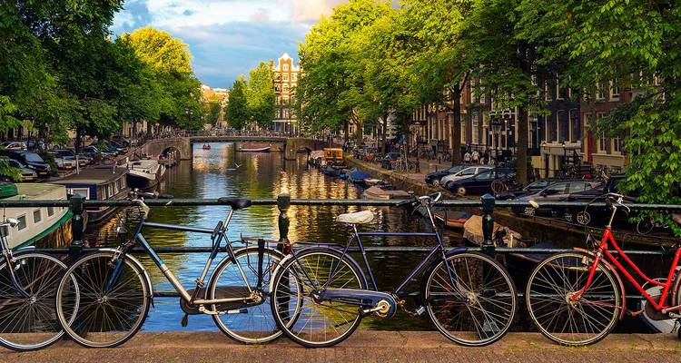 Des bicyclettes garées sur un pont surplombent un canal d'Amsterdam ensoleillé bordé de maisons historiques.