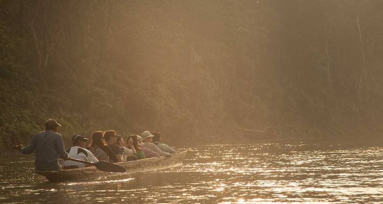 Kanu voller Reisender treibt bei Sonnenaufgang einen nebligen Dschungelfluss hinab, goldenes Licht filtert durch die Bäume.