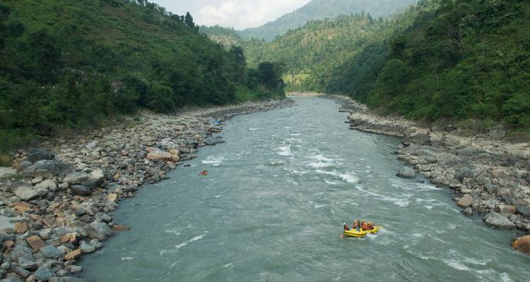 Ein aufblasbares Floß navigiert Wildwasser-Stromschnellen durch ein felsiges Tal eines Himalaya-Flusses, das von grünen Hügeln umgeben ist.