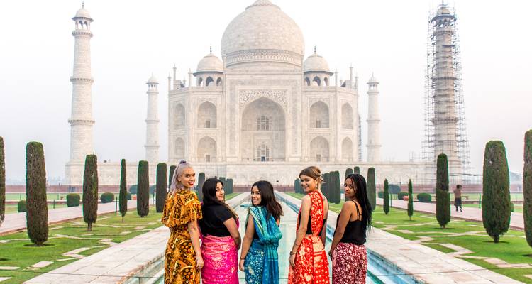 Un grupo de mujeres con coloridos saris posan frente al estanque de reflejos del Taj Mahal en una mañana brumosa.