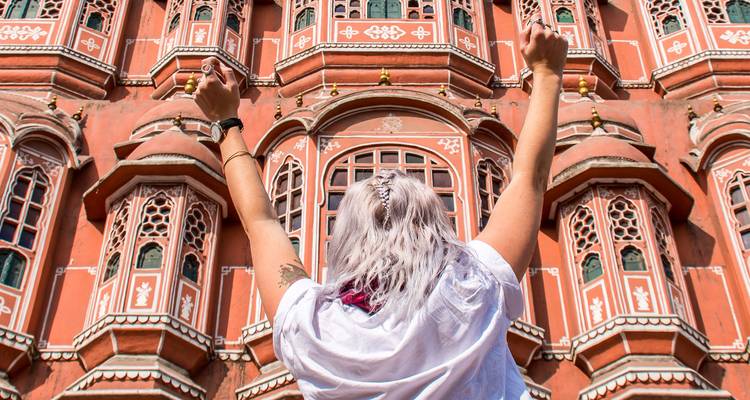 El viajero levanta los brazos en celebración frente a la ornada fachada rosa del Hawa Mahal de Jaipur.