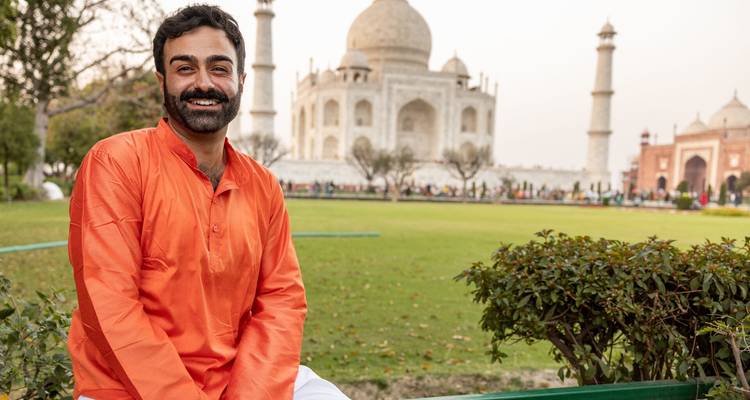Un hombre con kurta naranja se sienta en una baranda baja, sonriendo con los jardines del Taj Mahal y el mausoleo suavemente desenfocados de fondo.