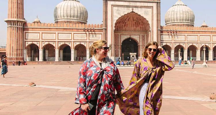 Twee lachende vrouwen in kleurrijke gewaden lopen over de immense binnenplaats van de Jama Masjid met zijn indrukwekkende koepels en bogen achter hen.