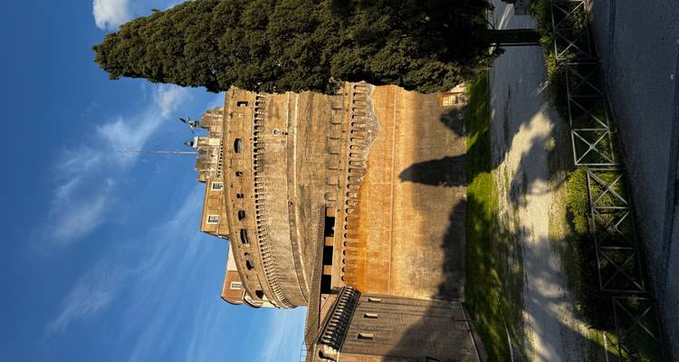 Massieve cilindrische muren van Castel Sant'Angelo steken uit boven de bomen onder een stralend blauwe hemel.