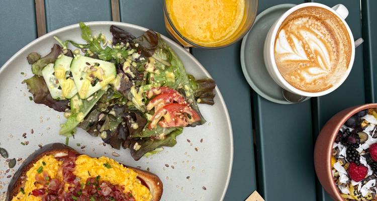 Vista aérea de un brunch con ensalada de aguacate, tostadas con queso, cappuccino, smoothie y yogur con frutos rojos sobre una mesa de patio.