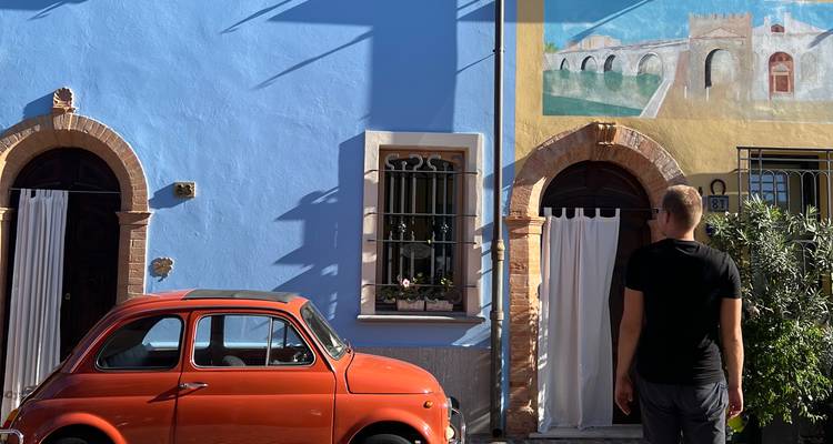 Scène de rue colorée avec une Fiat 500 rouge vintage garée contre un mur bleu vif et un homme admirant une fresque murale.