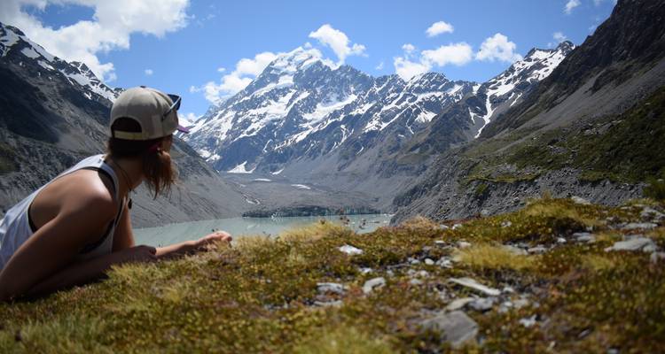 Un voyageur se détend sur l'herbe alpine en admirant un lac glaciaire turquoise et des pics enneigés.