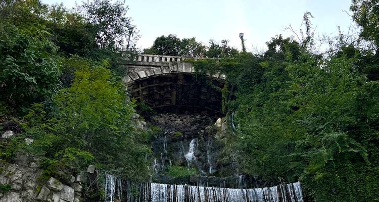 Kleiner kaskadierender Wasserfall, der unter einem Steinbogen fließt und von dichtem Grün umgeben ist.