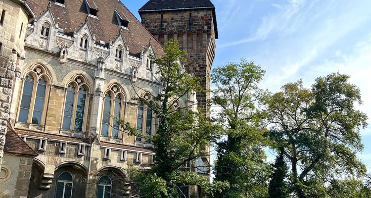 Historisches Stein- und Backsteingebäude mit gotischen Fenstern und hohem Turm neben belaubten Bäumen.