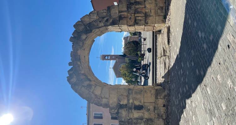 Une petite arche en pierre encadre une église et son clocher dans une place de ville ensoleillée.