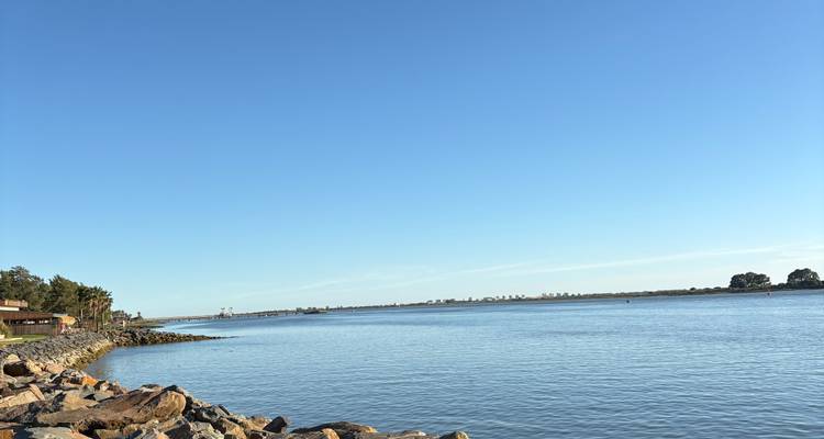 Scène de rivière calme bordée de rochers et d'une côte basse sous un ciel bleu clair.