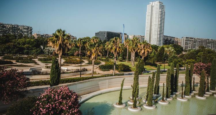 Vue sur un parc aménagé avec palmiers et cyprès donnant sur une tour résidentielle moderne de grande hauteur.