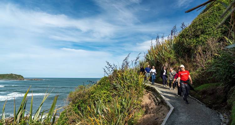 Kleine wandelgroep volgt een kustpad boven brullende golven en vlas struiken onder een blauwe hemel.