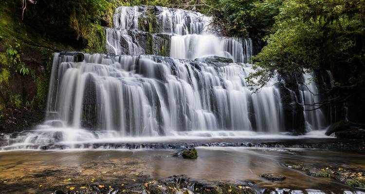 Zijdeachtige waterval met meerdere niveaus stort zich over mosbegroeid basalt af in een rustig bosmeertje.